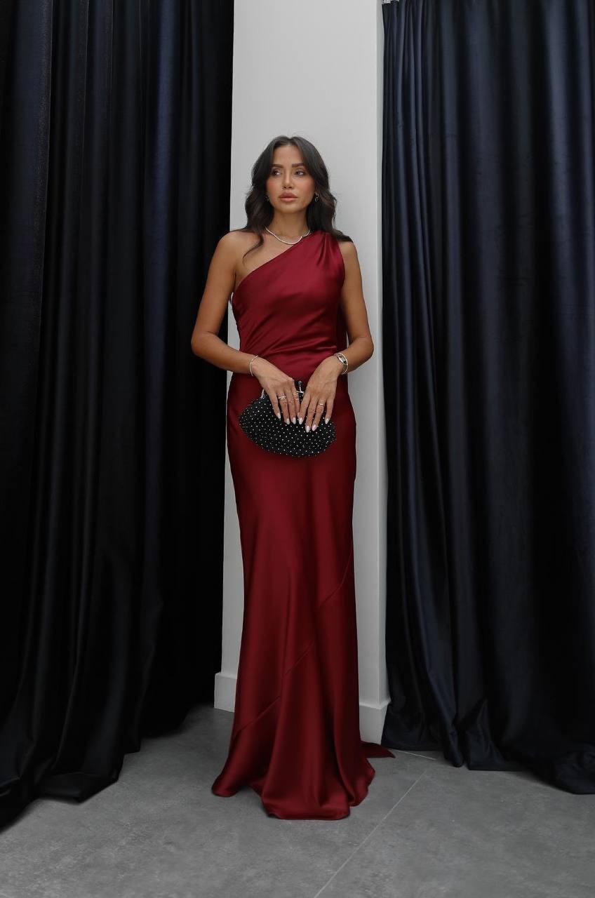Woman in a burgundy one-shoulder dress holding a black clutch against a black curtain background