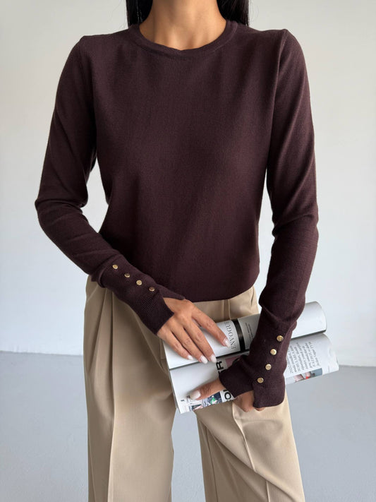 Person wearing a dark brown sweater with button details and beige pants, holding a book against a plain background.