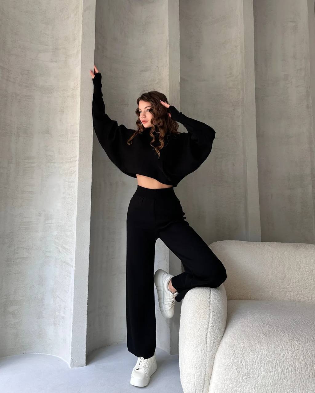 Person wearing a black outfit sitting on a white chair against a gray wall.