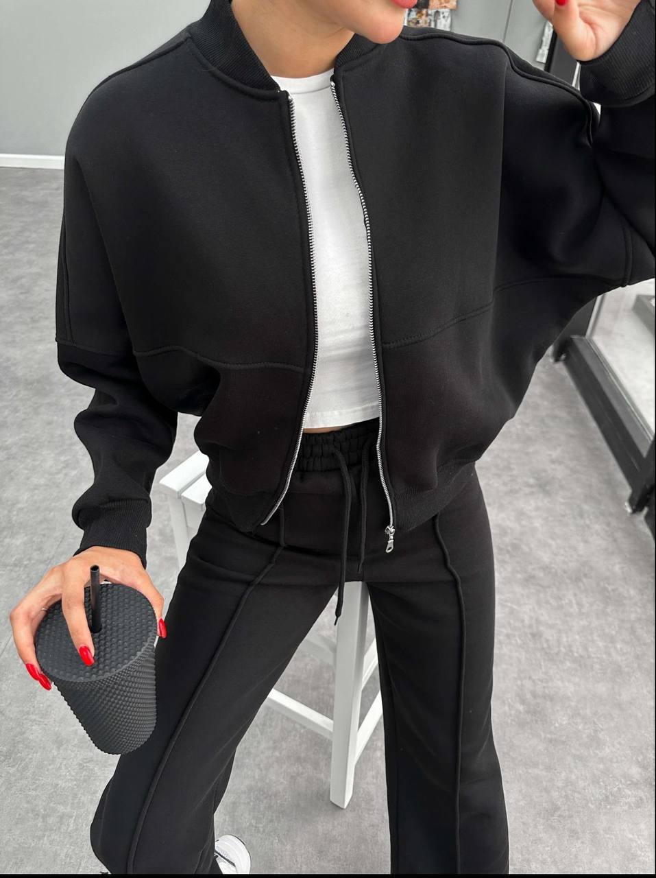 Person wearing a black tracksuit holding a black cup indoors.