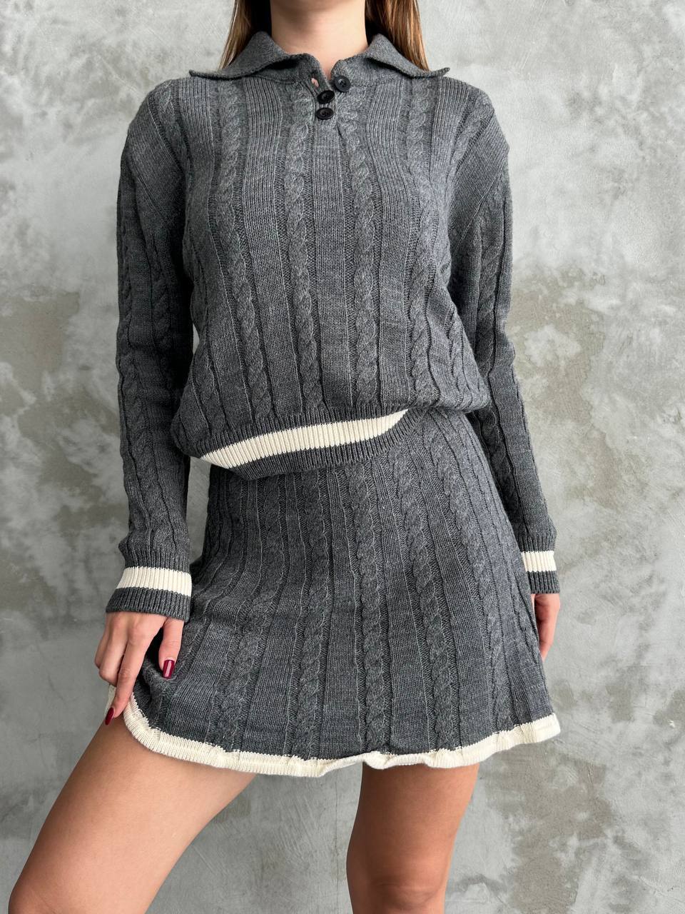 Person wearing a gray cable knit sweater and skirt set against a textured gray background