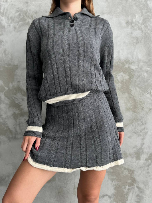 Person wearing a gray cable knit sweater and skirt set against a textured gray background