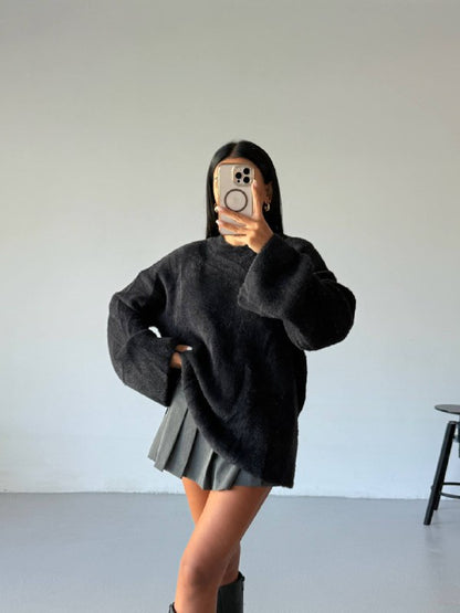Person wearing a black oversized sweater and pleated skirt in a minimalistic setting.