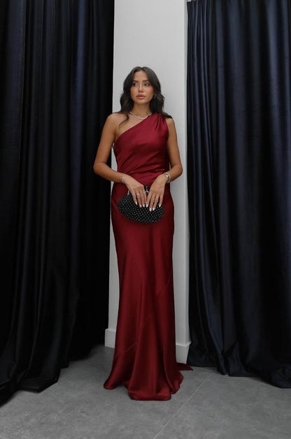 Woman in a burgundy one-shoulder dress holding a black clutch against a black curtain background