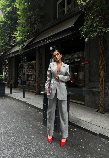 Woman in a gray suit with red shoes standing on a street in front of a store named 'MISELA'.