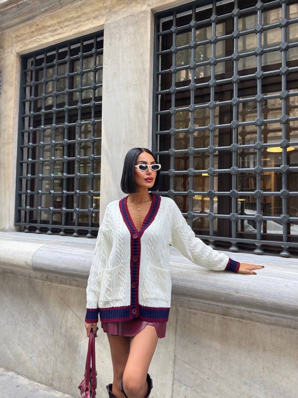 Woman in a white cardigan and purple skirt standing against a building with large windows.