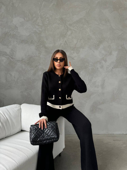 Woman in black outfit with sunglasses and handbag posing against a gray wall.