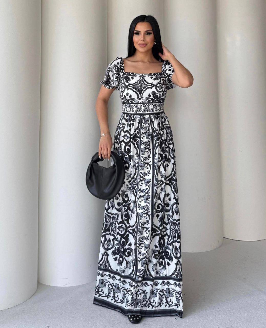 Woman wearing a black and white patterned dress holding a black handbag against a white curtain background