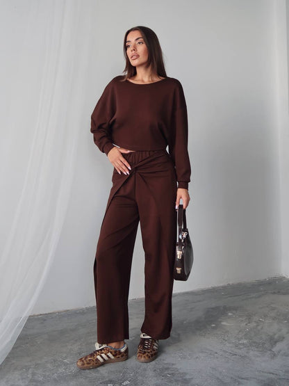Woman wearing a brown outfit holding a black handbag against a plain background