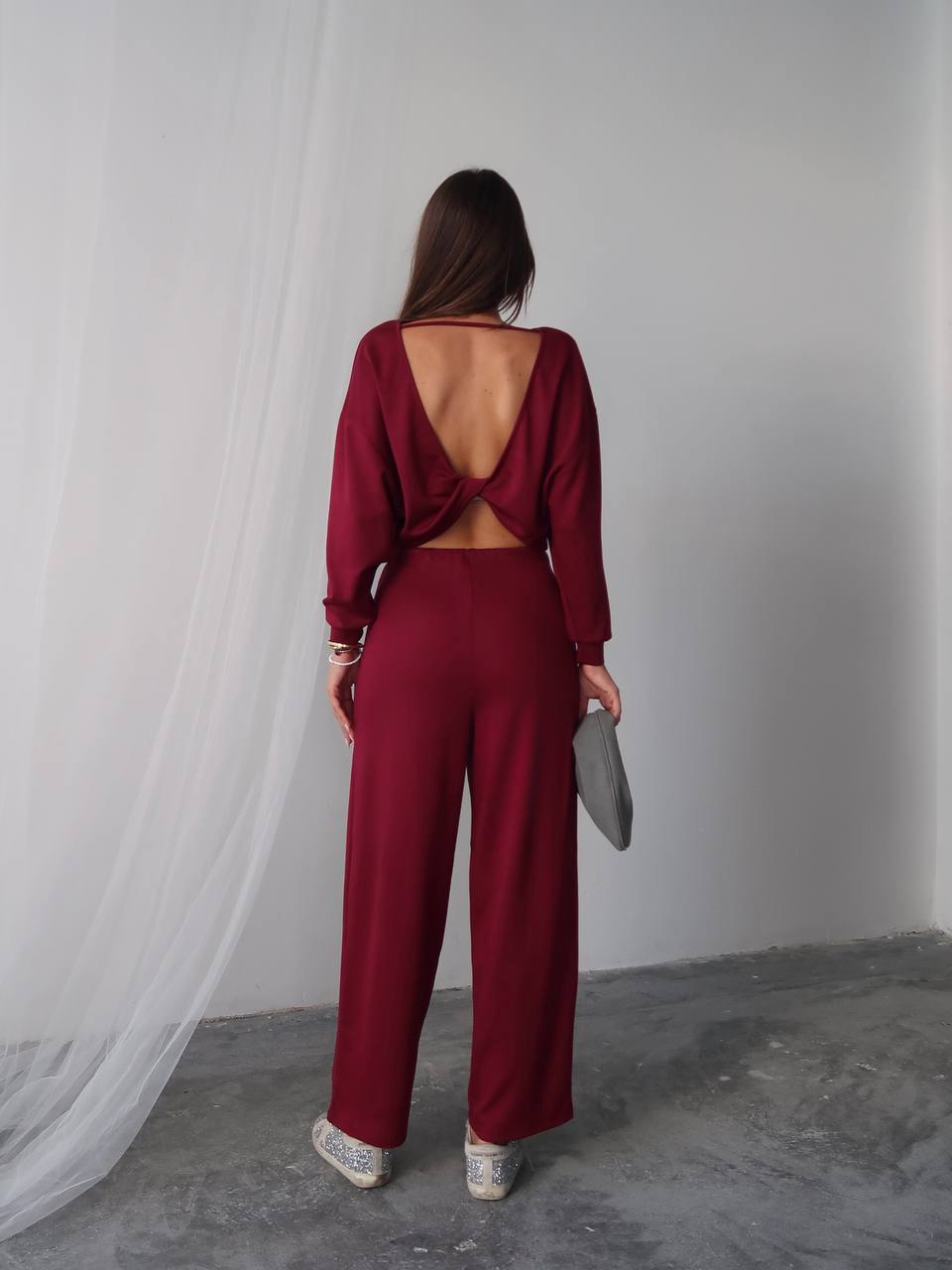 Woman wearing a burgundy open-back jumpsuit standing against a white curtain.