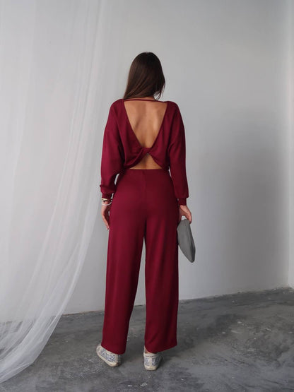 Woman wearing a burgundy open-back jumpsuit standing against a white curtain.