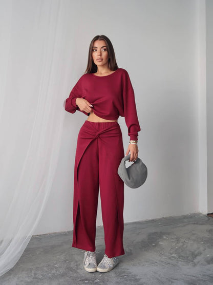Woman wearing a burgundy outfit with wide-leg pants and a matching top, holding a gray clutch against a white curtain background.