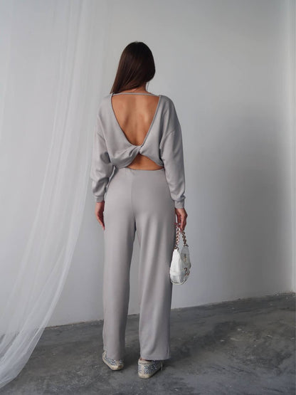 Woman wearing a light gray open-back top and high-waisted pants against a white wall.