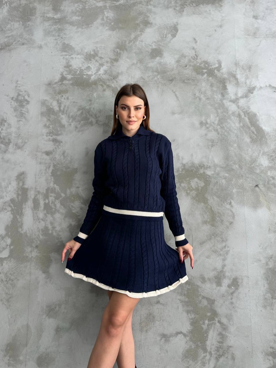 Woman wearing a navy blue cable knit sweater and skirt with white trim against a textured gray wall.