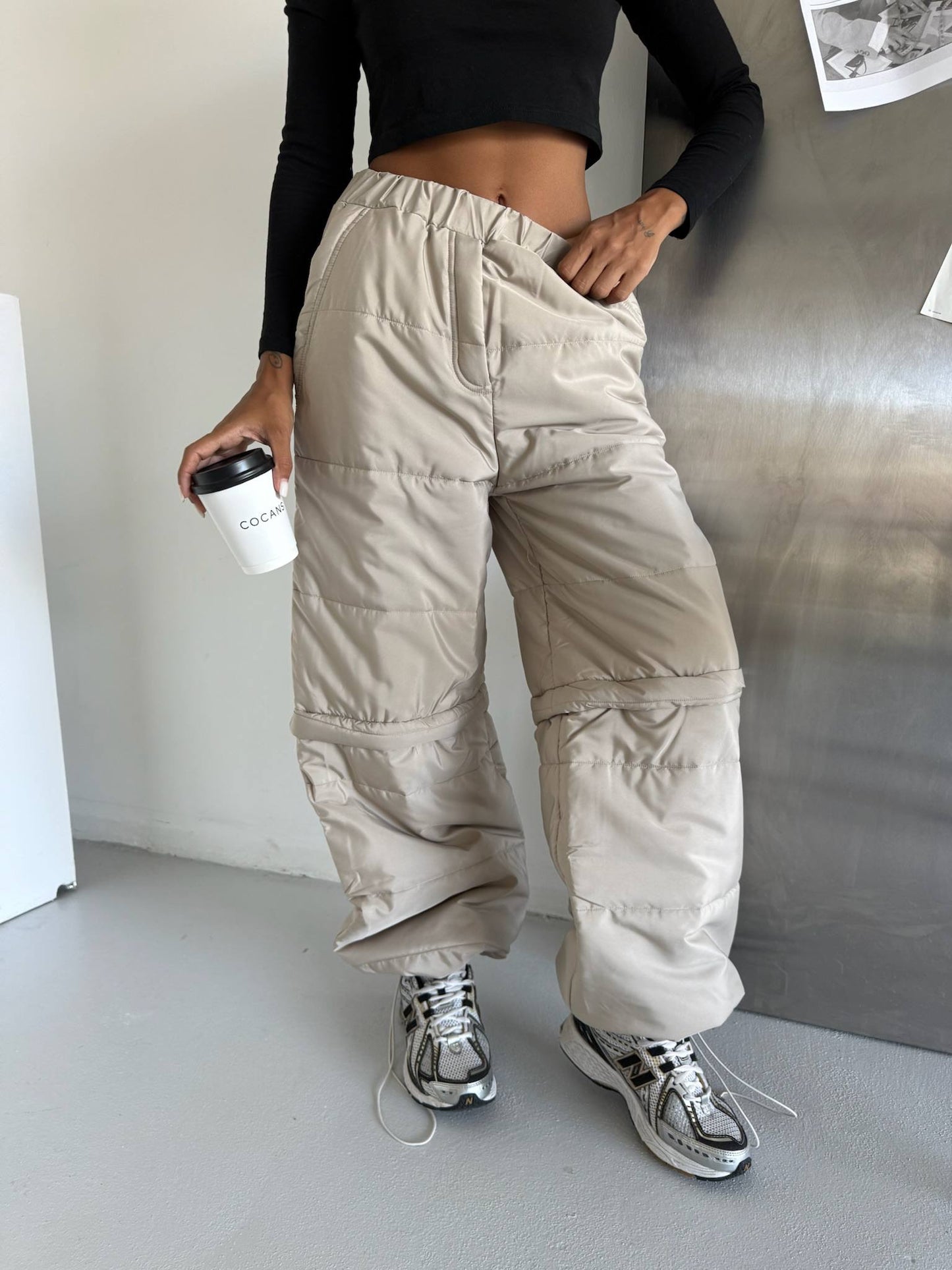 Person wearing beige cargo pants and black top, holding a coffee cup indoors.
