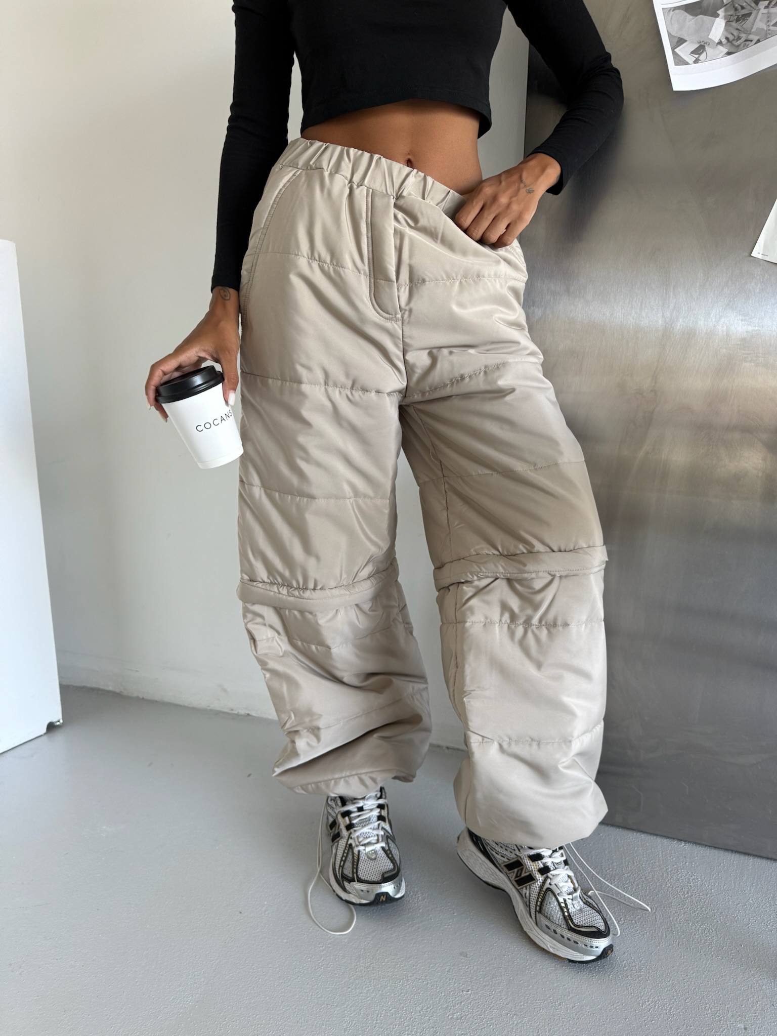 Person wearing beige cargo pants and black top, holding a coffee cup indoors.